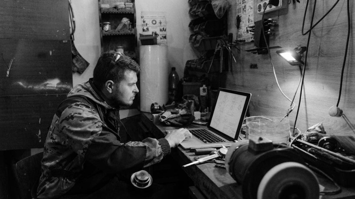 Black and white photo of a craftsman working on a laptop in a workshop setting.